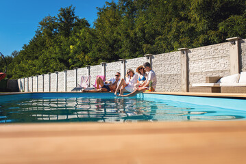 Group of adult friends sitting and relaxing next to outdoor pool during summer vacation.