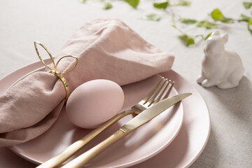 Easter table setting with pastel pink egg, gold cutlery and linen napkin on soft pink plate. Elegant spring decor.
