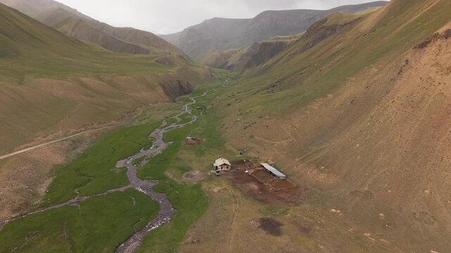 Kyrgyzstan - A Small Homestead Sits in a Wide Green Valley, Surrounded by Rugged Hills and a Winding Stream Flowing through the Landscape - Aerial Drone Shot