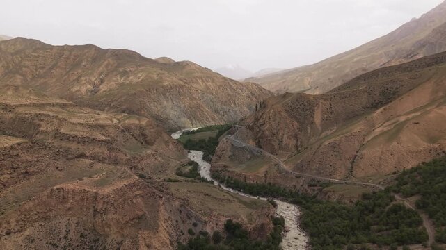 Kyrgyzstan - A Winding River Flows through Rugged, Brown Mountain Valleys Dotted with Patches of Green Trees - Aerial Drone Shot