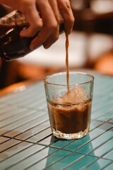 Hand pouring chilled coffee concentrate into a glass of ice. Close-up beverage preparation in a warm café setting, artisan coffee craft, lifestyle drink culture and hospitality workflow.