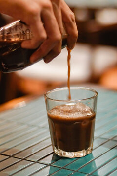 Close-up of a barista pouring iced coffee concentrate into a glass filled with ice cubes. Beverage preparation moment, warm caf&eacute; ambience, artisan drink craft and lifestyle coffee culture scene.