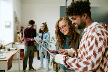Young adult Black man and young adult Caucasian woman using digital tablet together in modern office while two young adult colleagues collaborating in background, all smiling and interacting