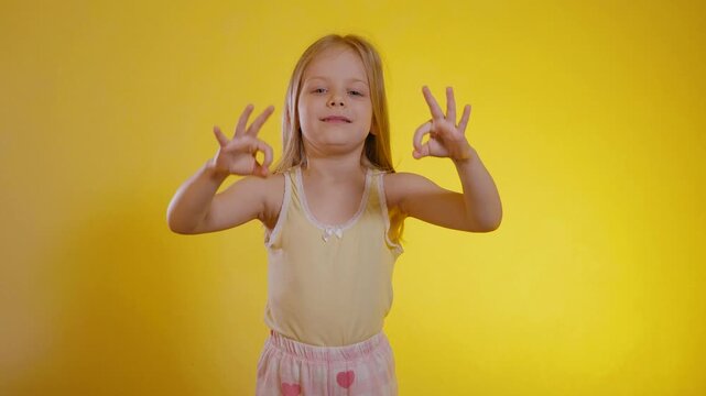 Young girl with blonde hair in a yellow top making the 'okay' gesture on both sides against a vibrant yellow background. Close-up of a cheerful kid displaying 'A-OK' hand signs, illuminated by warm 