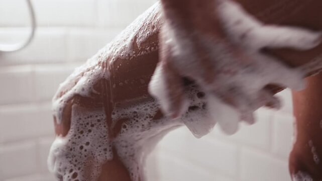 Close-up of a womans leg being lathered with soap in a shower, showcasing the cleansing process, camera zooms in and captures the texture of the foam and skin