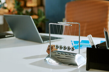 Closeup showing Newtons cradle swinging on modern office desk with open laptop and documents in background, illustrating concept of physics, motion, and business productivity