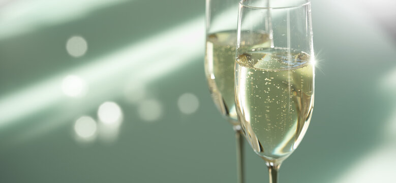 Two filled champagne glasses on a modern glass table. Elegant modern style with soft bright green background und light bokeh. Horizontal close-up.