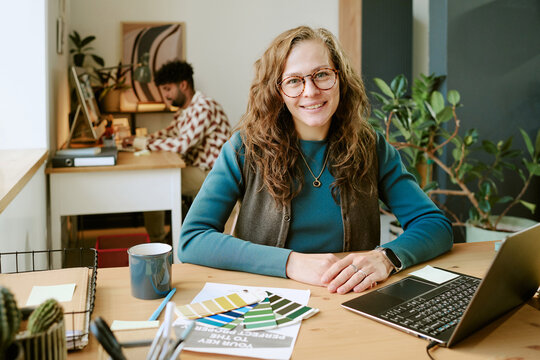 Portrait of middle aged Caucasian woman with long curly hair smiling at camera while sitting at desk with laptop and color swatches, young Black man working in background - Powered by Adobe