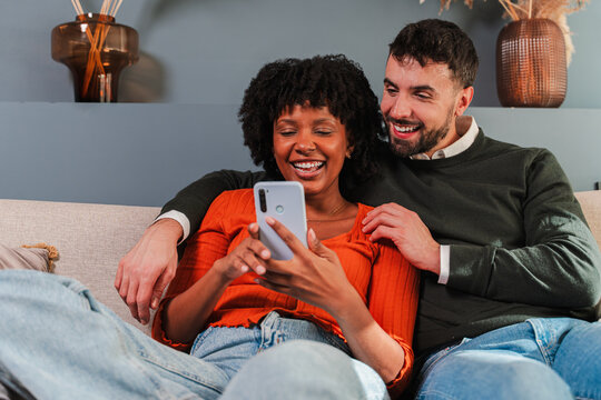 Delighted couple enjoying their time together while interacting with a smartphone on a comfortable couch, sharing laughter and joy in a cozy setting filled with love and connection. - Powered by Adobe