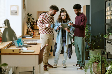 Three young adults, two Black men and one Caucasian woman, standing together in modern office, collaborating while using digital tablet, discussing project near desk with laptop