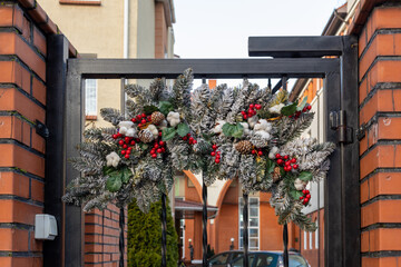 Frosted fir branch Christmas garland with red berries, pinecones, cotton and green leaves on black metal gate. Outdoor entrance decoration.
