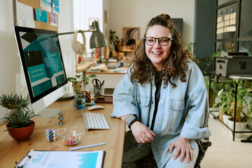 Portrait of young adult Caucasian woman with long curly hair and glasses sitting at desk in modern office smiling at camera, computer monitor and office supplies visible on workspace