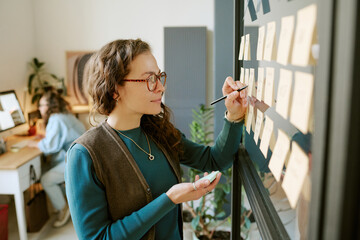 Caucasian young adult woman writing on sticky notes attached to glass wall, holding marker and eraser, working in modern office while another young adult woman works at computer in background