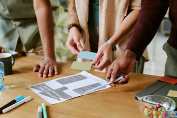 Multiethnic group of young adult men and women collaborating around table, holding color swatches and discussing printed document, hands and arms visible, working on creative project