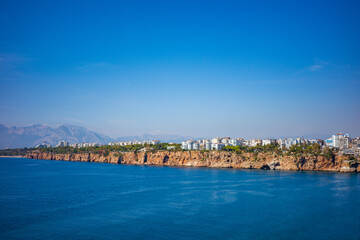 Obraz premium A vibrant panoramic view of Antalya’s rocky coastal cliffs meeting the deep blue Mediterranean Sea, with modern white apartment buildings lining the shoreline and mountains
