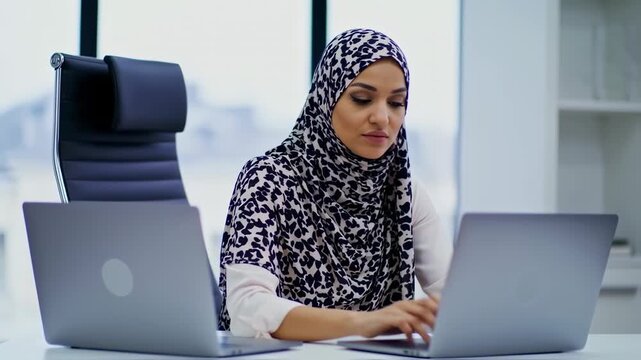 Woman in patterned headscarf types on laptop at a desk, two laptops present in office