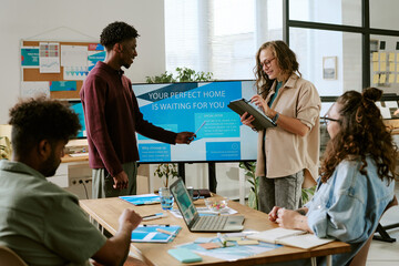 Diverse group of young adult men and women collaborating in modern office, Black man presenting information on digital screen while Caucasian woman taking notes on tablet, others listening