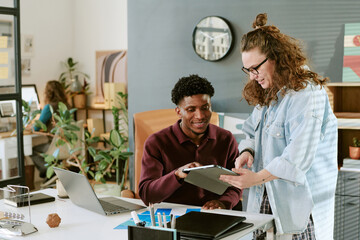 Young adult Caucasian woman showing digital tablet to young adult Black man sitting at desk, both collaborating on project in modern office environment with plants and artwork visible
