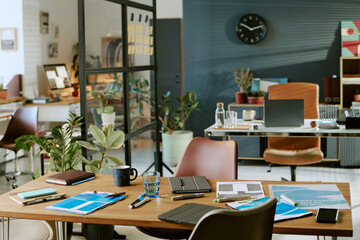 Modern office workspace showing empty chairs around table with documents, notebooks, pens, glass of water, digital tablet, laptop and potted plants in background