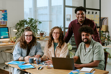 Group of young adult multiethnic men and women sitting at desk working together on project, smiling at camera, using laptop and documents in modern office setting