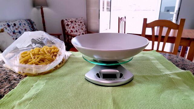 Home scene of a person measuring the correct portion of penne pasta, a staple of Italian cuisine and the Mediterranean diet, pouring it from a bag into a bowl on a digital kitchen scale