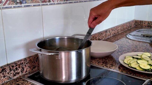 Home cooking scene shows a person's hand stirring boiling water in a large steel pot on a modern stove, preparing a healthy meal with fresh sliced zucchini on the side