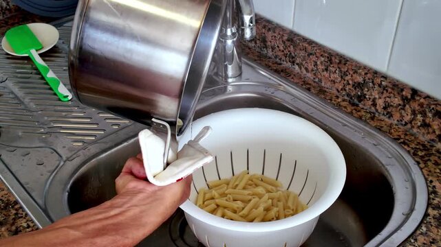 Unrecognizable person draining hot water from boiled penne pasta in a white colander over a kitchen sink, preparing a typical Italian meal that's part of the Mediterranean diet