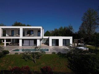 Modern luxury villa with pool, terraces, and clean minimalist architecture captured by drone. Bright sunny day, surrounding greenery, and contemporary outdoor living areas