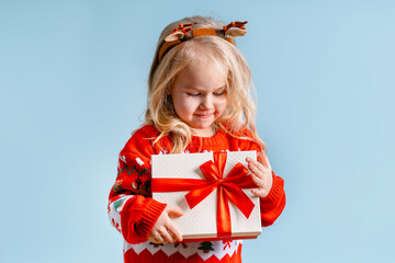 Portrait of a cute little child girl in christmas sweater with a deer antlers rim on an isolated blue background. Holds a gift box. Preparing for the New Year holidays and Christmas