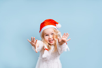 Obraz premium Cheerful little child girl in a white dress and Santa hat on a blue background, the concept of new year and Christmas, space for text on the banner