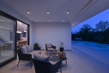 Exterior view of a modern luxury house with a swimming pool at twilight. Illuminated two-story white villa with large glass windows, patio, terrace, and glowing blue water