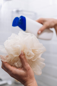 Close-up of hands holding a soft shower loofah while applying liquid soap in a bright bathroom, showcasing personal care and hygiene routine with copy space