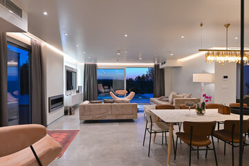 Modern luxury living room interior at sunset. Open-plan space featuring beige sofas, marble dining table, large glass windows with sun flare, and view of a patio. Stylish contemporary home design