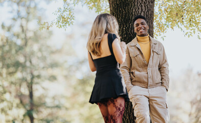 Fototapeta premium Two friends share a friendly moment in a sunlit park. One wears a beige jacket and mustard turtleneck, the other a black dress. A candid outdoor scene conveying warmth, connection, and casual style.