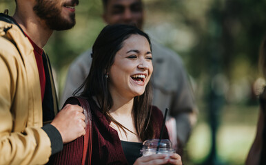 A group of friends share a joyful moment in a park, laughing and chatting while one holds a drink. Bright daylight and greenery create a lively, carefree outdoor scene.