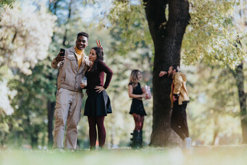 A diverse group of friends enjoys a sunny day in the park. Foreground couple selfies while others chat nearby, capturing casual, joyful moments in nature.