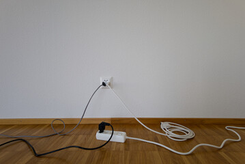 Tangled power cords and an extension strip on a wooden floor. Minimalist scene with a wall socket, cables, and plug connections. Swiss power socket. Home electricity setup and consumption.