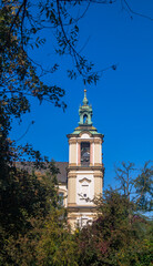 Church bell tower among leaves and tree branches. Old bell tower with green roof against blue sky