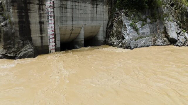 Aerial drone view of a concrete hydropower intake structure on the muddy banks of a flowing river in the lush green mountains of Nepal for renewable energy production project