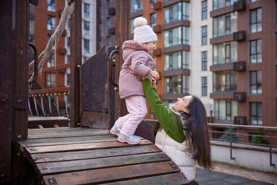 Happy mother raising up daughter on playground with city buildings in background - Powered by Adobe
