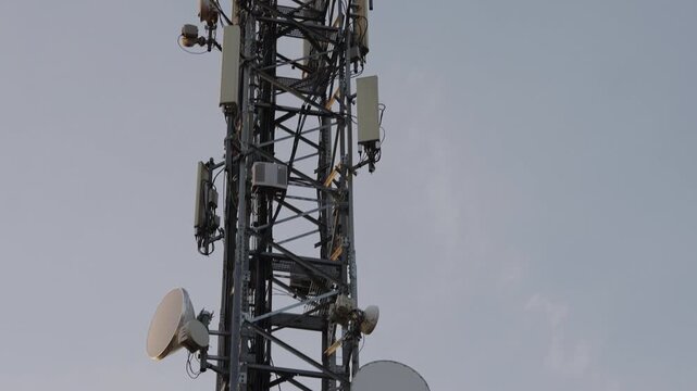Telecom tower with antennas and dishes, steel lattice rising above tree line at dusk, closeup on transmitter arrays and microwave panels, maintenance crew