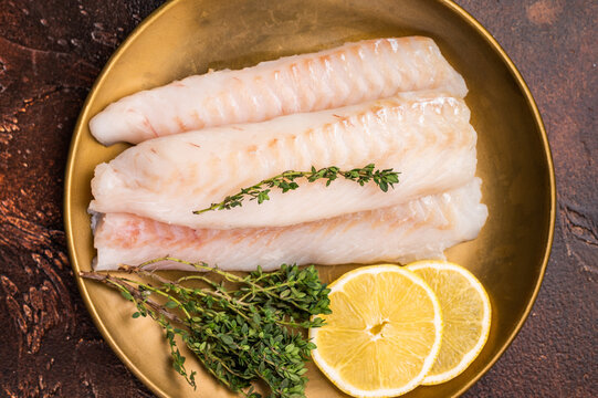 Raw cod loin pieces, whitefish fillets without skin on plate with herbs. brown background. top view