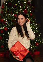 Woman in a Christmas decorated room poses in front of a tree, holding a red gift with ribbon and smiling. She places a large bow on the Christmas tree, festive interior, holiday mood.
