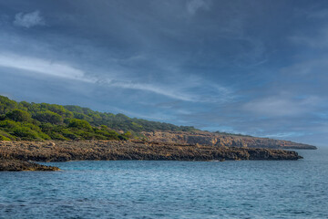Rugged rocky Mediterranean coastline and dark blue sea with green forest under a cloudy blue sky at sunset with copy space