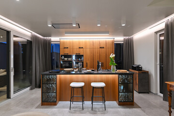 Modern luxury kitchen interior with wooden cabinetry, central island with bar stools, and ambient LED lighting. Stylish contemporary design with large windows and dark countertops at twilight