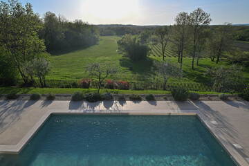 Fototapeta premium Elevated view of a modern swimming pool with clear blue water, overlooking a picturesque, expansive green landscape of fields, trees, and vineyards under a bright sky. Perfect for tranquil luxury