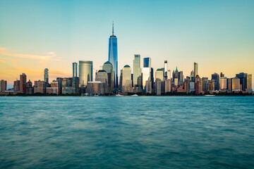 Obraz premium The iconic Downtown Manhattan skyline shines brightly during the blue hour, viewed across the smooth waters of the Hudson River.