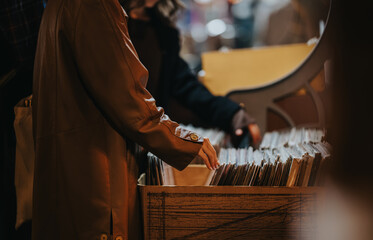 A person in a tan coat reaches into a crate of vinyl records at a market stall, with warm lighting...