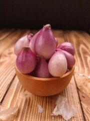 A small wooden bowl filled with red onions, sitting on a wooden surface. The onions are bulbous with a reddish-purple hue and smooth skin.