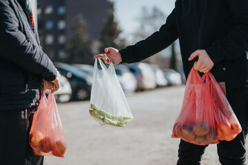 Two individuals share a moment of shopping on a city street, passing plastic bags with groceries. The scene captures daily life, casual interaction, and simple, communal moments.